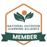 Image of the badge for members of the National Outdoor Learning Alliance. The badge is a brown hexagon outline turned so a point is facing directly up. A green banner across the bottom of the hexagon has the word "member" on it in white print. In the center of the hexagon green lettering reads "national outdoor learning alliance" and above those words is the logo image of four green leaves, two layered on the left and two layered on the right, with a brown circle above them in the center. This badge indicates that the business which posted it is a member of NOLA.